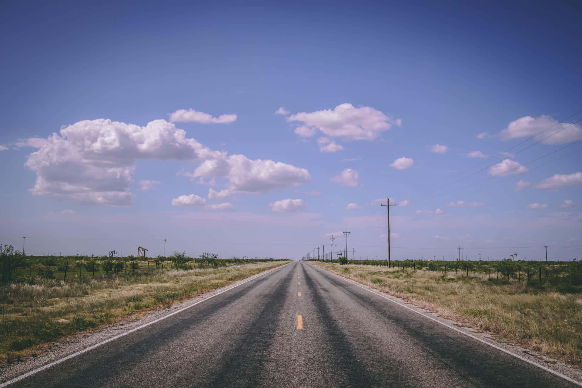 A long, straight road stretches through the rural landscape of Odessa, TX, under a bright, clear sky.